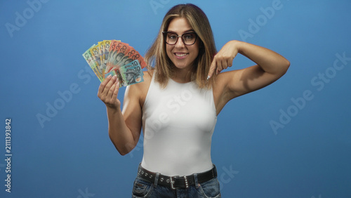Fotografie Young hispanic blonde woman wearing white sleeveless top and glasses holds a fan of australian banknotes and points finger to cash in studio; confidence wealth