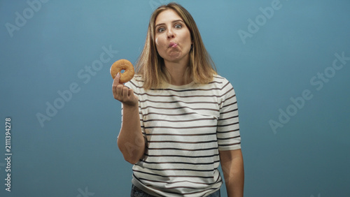 Foto Woman holding donut with fingertip and sticking tongue out while looking at camera against blue studio wall in a striped shirt; playful curiosity