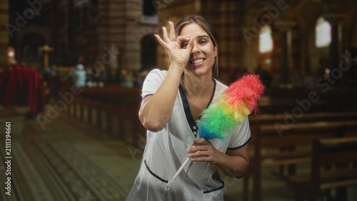 Obraz na plátně Woman cleaner in white uniform holding a rainbow feather duster makes ok sign to eye inside an ornate cathedral aisle with wooden pews and stained glass windows visible; playful
