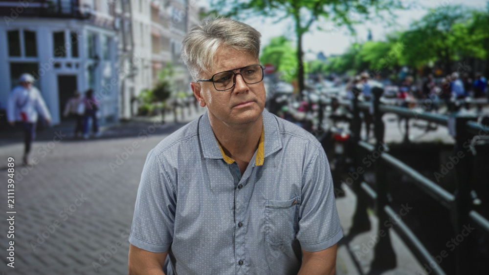 Fototapeta premium Man wearing glasses standing on a busy street by a canal railing, looking sideways and holding neutral expression; pensive reflection.