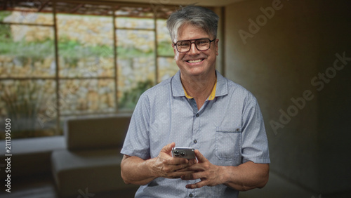 Foto Senior man holding smartphone and smiling with visible hands in a building lounge by a large window and sofa; friendly contentment