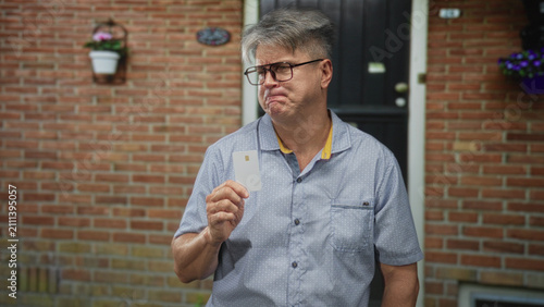 Obraz na płótnie Man holds creditcard in hand before brick building doorway, frowning with glasses and pursed lips; disappointment money concern