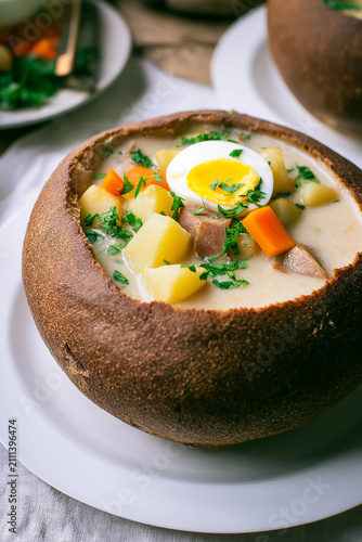 Polish sour rye soup with sausage, potatoes, carrots and a halved egg served in a bread bowl. Comfort food, traditional cuisine, rustic dining, hearty meal, cozy lunch, farmhouse cooking.