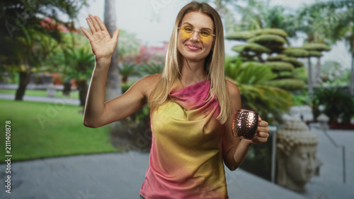 Photography Blonde woman holding a copper cocktail mug waves hand toward camera in building courtyard, wearing yellow sunglasses and gradient dress; cheerful celebration