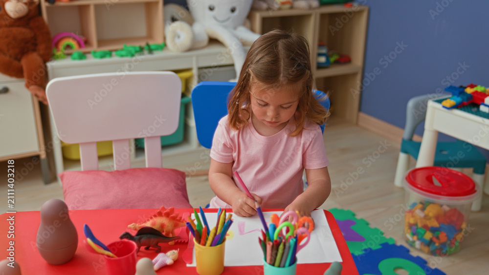 Fototapeta premium Little hispanic girl draws with crayon on paper, reaching hand across a red table in a playroom building; creative concentration.