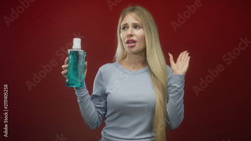 Foto Woman young blonde holding mouthwash bottle with eyes closed and grimacing, right hand raised in studio; disgust aversion oralcare