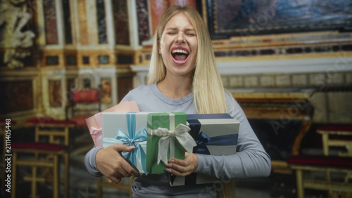 Fotografie Woman holding several wrapped gifts, mouth open in a loud scream in a building ornate hall; excitement celebration