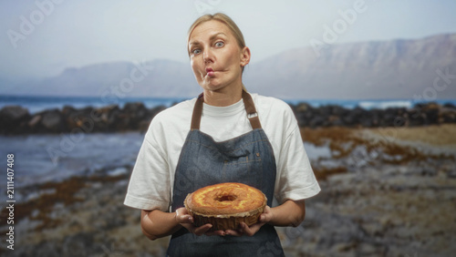 Bild auf Leinwand Woman middleaged blonde in apron holding sponge cake with puckered lips on a studio set with coastal backdrop; playful delight