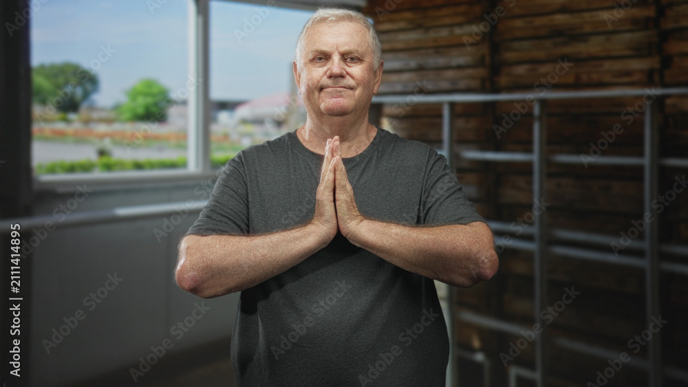 Fototapeta premium Senior man stands with palms pressed together in prayer gesture in a studio by a window and wooden wall, wearing a grey tshirt; calm reflection.