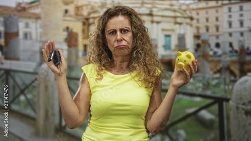 Fotografie Woman holding car keys in one hand and yellow piggy bank in the other, hands raised and frowning in front of building; financial dilemma frustration