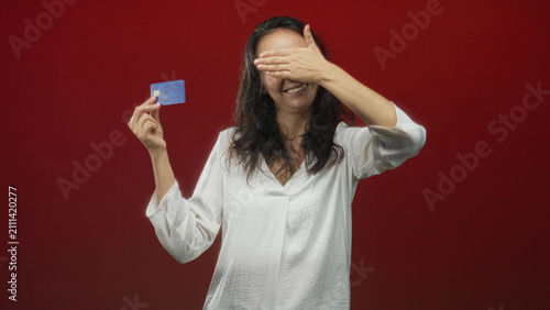 Фотография Woman holding creditcard and smiling covers eyes with hand while presenting a blue chip card against a red studio backdrop; playful secrecy