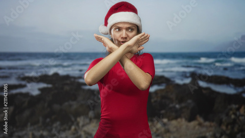Fotografie Woman in santa hat and red shirt crosses arms and gazes toward waves on a rocky shore set in a studio; holiday melancholy