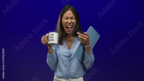 Canvastavla Young thai woman holds mug reading i am the boss and blue clipboard, mouth open and eyes closed while screaming in studio; leadership frustration