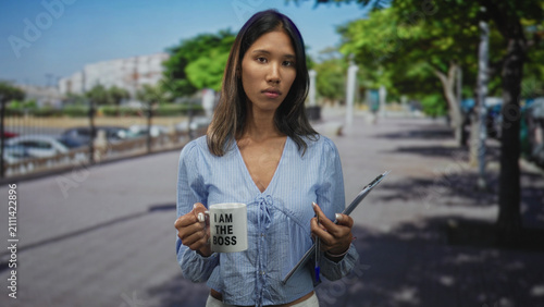 Konstfotografi Young thai woman holds a mug reading i am the boss and a clipboard, hand gripping mug and pen visible, standing on a busy city street; confidence leadership
