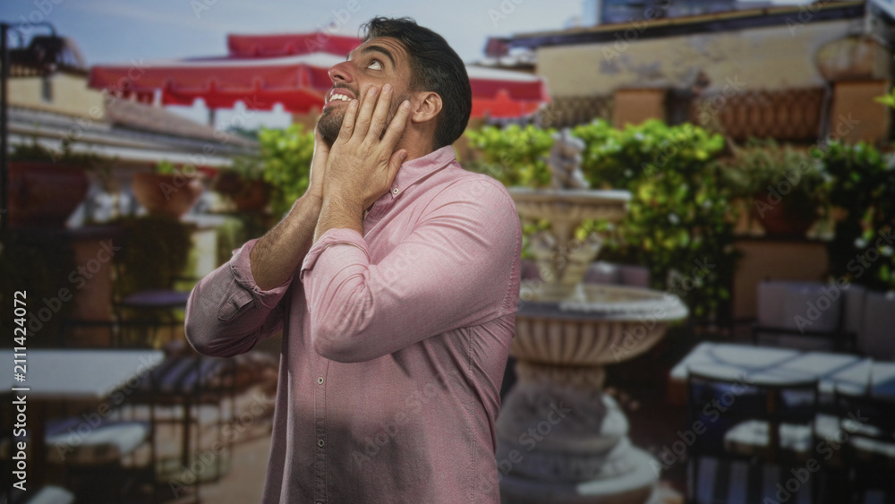 Fototapeta premium Man with hands covering cheeks looking upward on building terrace beside stone fountain, wearing pink shirt; surprise contemplation.