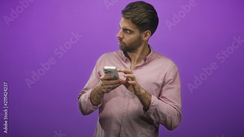 Cuadro en lienzo Bearded man holding smartphone, fingers tapping while glancing sideways, wearing pink shirt against purple studio backdrop; focus curiosity