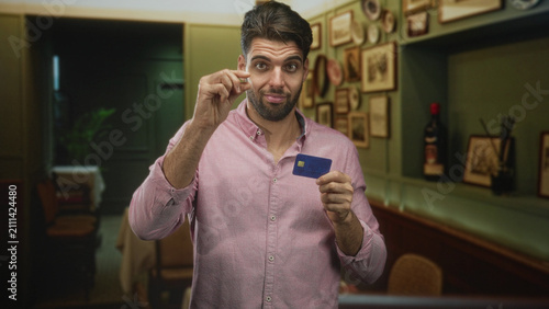Foto Man holding creditcard between hands inside a restaurant building, shows card and shrugs with beard and pink shirt; confidence convenience