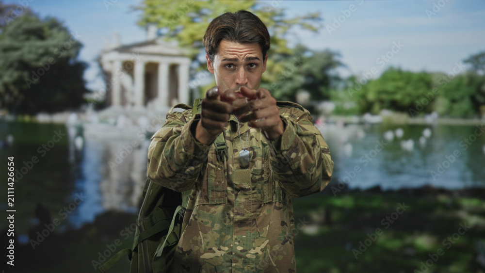 Fototapeta premium Man soldier in camouflage with hands clasped then open palms and pointing, tongue out, wearing dogtags and backpack near a classical building by a lake; playful camaraderie.
