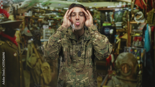Man soldier in camouflage touching temples with hands, dog tags visible, wearing uniform inside a military surplus shop crowded with gear; pride duty.