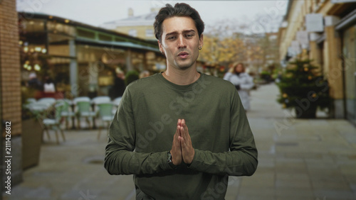фотографія Man with hands pressed together in pleading gesture on street cafe walkway wearing green shirt and watch, palms visible; regret plea