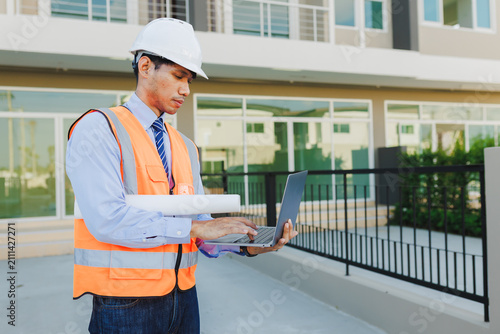 Male property inspector or engineer in high-visibility vest and hard hat, checking data on laptop computer near a new townhouse building.
