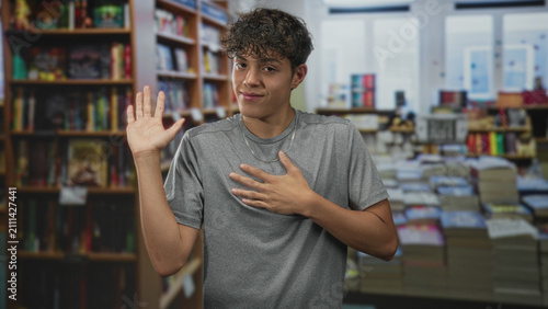 Принти на полотні Teenage boy with palm raised and hand on chest wearing gray shirt and silver chain in a bookstore building; sincerity promise