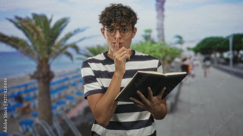 Fototapeta premium Teen boy in striped shirt and round glasses holds an open notebook and places finger to lips in a shushing gesture on a palm lined street by the beach; quiet mischief.