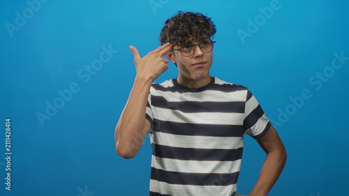 Quadro su tela Boy showing three finger hand flick with bare hand near face, wearing round glasses and horizontal striped shirt in studio against a blue backdrop; thoughtful curiosity