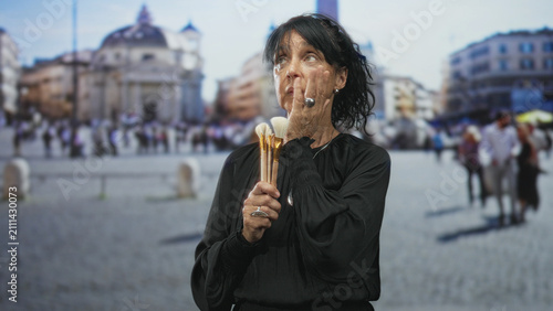 Foto Woman holding makeup brushes with hand on chin in street, city square visible behind her; contemplation creativity