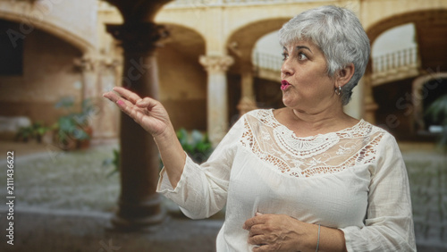 фотографія Woman pointing finger, hand on chest, speaking, wearing white lace blouse and hoop earring in a historic building courtyard; curiosity memory