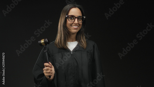 Фотография Woman judge wearing glasses and black robe stands holding a gavel in her right hand smiling in studio; confidence authority