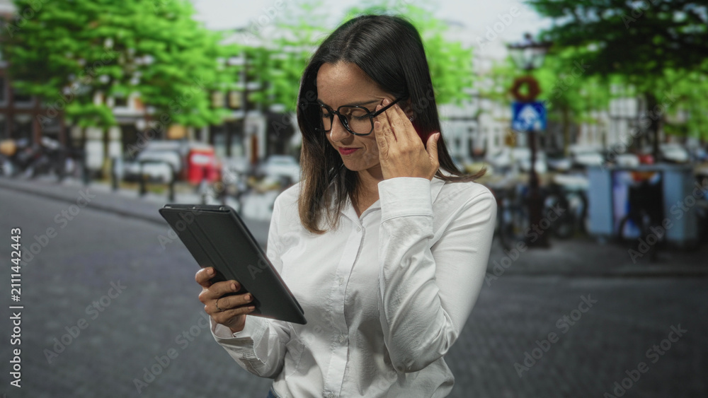 Fototapeta premium Woman in white shirt holding a tablet with one hand and touching glasses at her temple while reading on a street; concentration study.