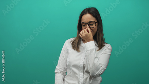 Canvastavla Woman with glasses pinches nose with hand in studio wearing white shirt and smiling near a teal wall backdrop; amused