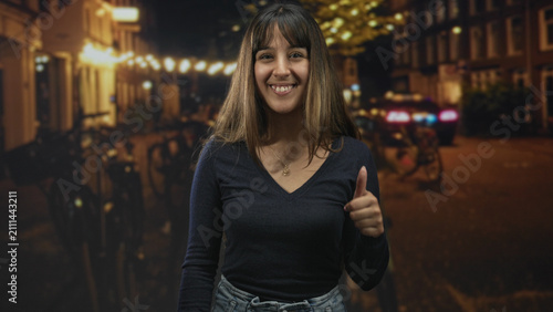 Fotografía Woman giving thumbs up on a night street lined with bicycles and warm string lights; joy confidence