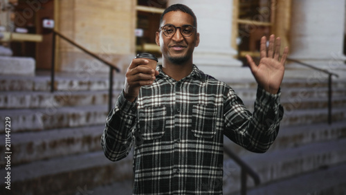 Quadro su tela Young black man wearing glasses and plaid shirt holding a takeaway coffee cup and waving right hand at a stone building entrance with steps; friendly greeting