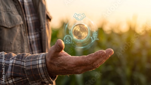 Agricultural investment concept, farmer holding glowing coin with handshake, barn, wheat, checkmark icons in field at sunset, farm funding, agribusiness deal and rural economy