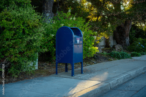 Fotografie Classic blue mailbox stands on a sidewalk next to lush green bushes