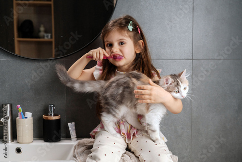 Girl brushes teeth while holding a cat in a bathroom setting in the morning