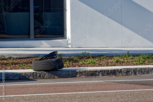 A discarded car tire and detached rim sit on the curb beside a modern building, casting long shadows on the sunlit sidewalk and highlighting urban neglect, roadside waste and potential safety hazards.