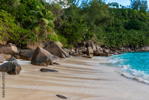 Canvastavla Quiet sandy shoreline framed by lush jungle and granite rocks beside clear blue water on a sunny day in the Seychelles