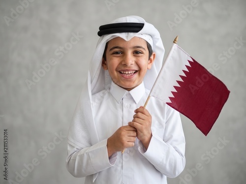 Smiling young boy wearing traditional white thobe and ghutra holding a small qatar flag against a neutral grey textured wall background