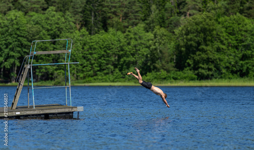 Young man finishing headfirst dive beside wooden platform on blue lake.