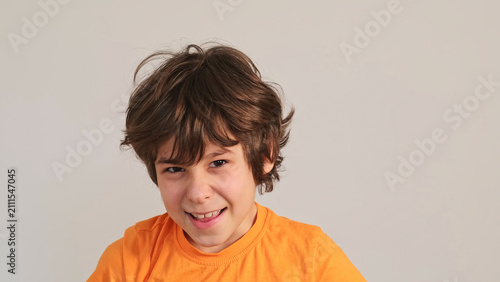 Close portrait of a smiling boy in an orange shirt making a playful face against a plain wall with copy space. Childhood emotions, confidence, and simple indoor play support family lifestyle stories.