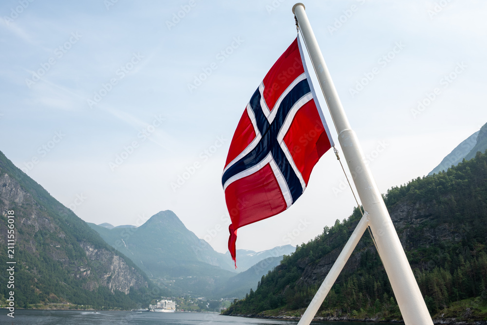 Fototapeta premium Red Norwegian flag waving on a boat with mountains and fjord water in the background on a bright summer day.