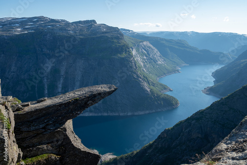 Dramatic cliff ledge above a deep Norwegian fjord valley under clear blue sky, showcasing rugged mountains and glacial lake scenery.