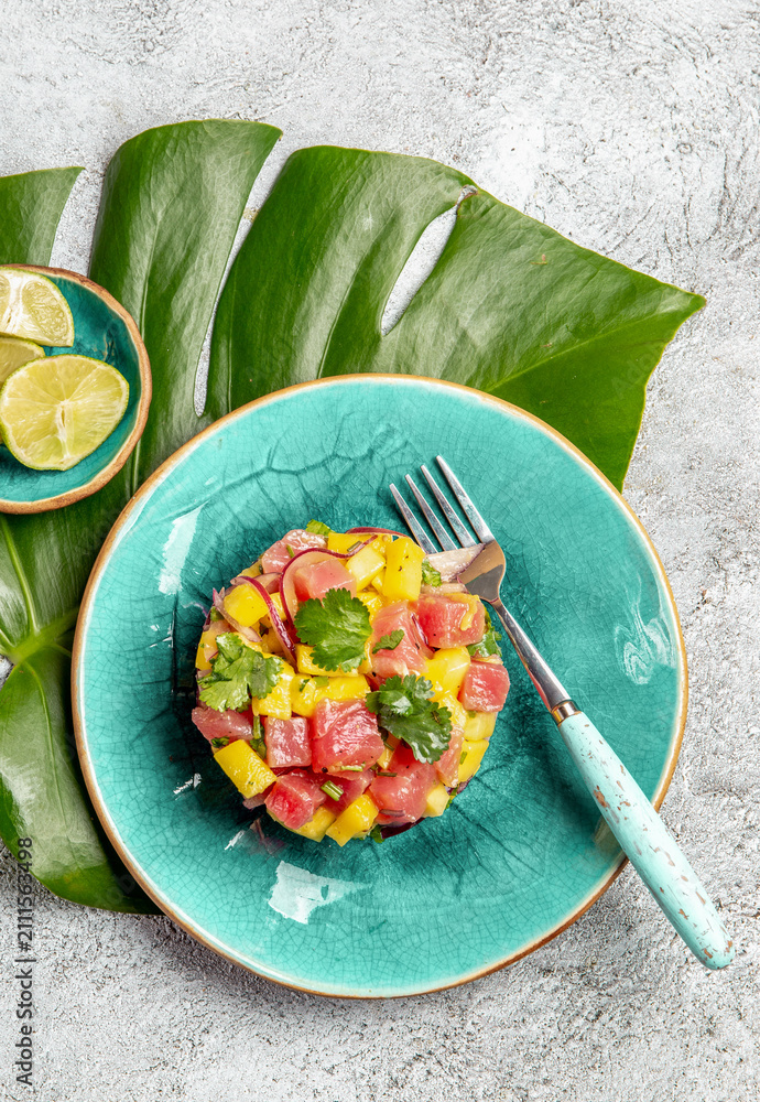 Obraz premium Tropical style gourmet tuna mango salad tartar with cilantro and purple onion. On blue plate, tropical leaves