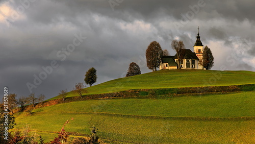 Fotografie Sorrowful Mother of God church (Cerkev Žalostne Matere Božje) on a hilltop of the Prealps in a cloudy but sunlit, autumn dawn