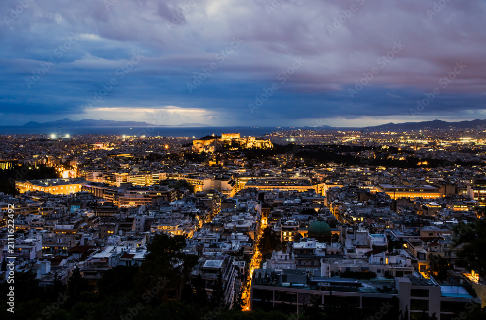 Obraz premium Panoramic night view of Athens city, Greece with the illuminated Acropolis and Parthenon visible on the hill. City lights, the sea in the background, and the iconic ancient monument