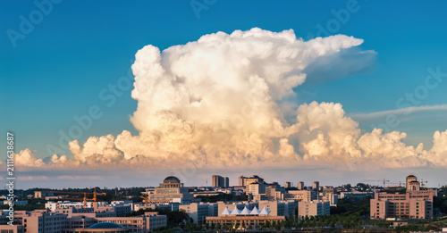 Bild auf Leinwand A massive cumulus cloud looms over a sprawling urban cityscape during sunset, with clear blue sky contrasting warm cloud tones