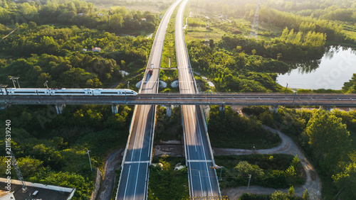 Fotografie An aerial view of a high-speed train traveling on an overpass crossing a highway through lush green landscape with water bodies